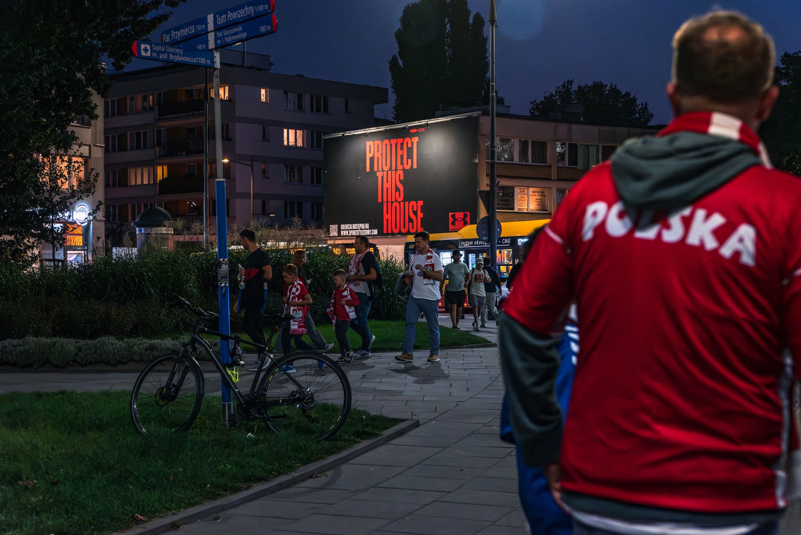 szeroki kadr przedstawiający kibiców idących na mecz Polski, na drugim planie mural dla marki Under Armour, duże litery na czarnym tle "Protect This House" 
